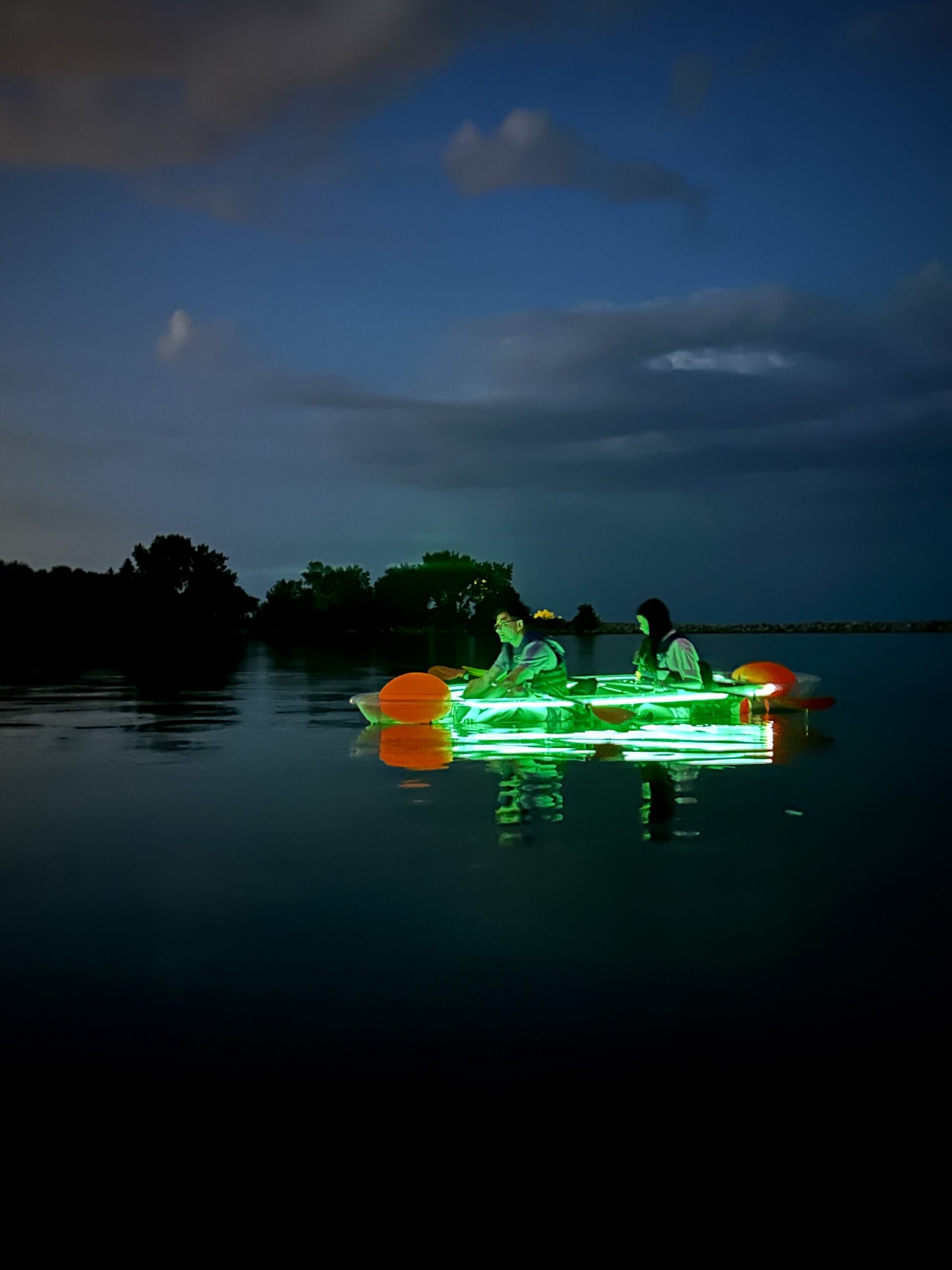 Couple in a glowing green LED clear kayak paddling at night on calm water in Toronto - a unique glow in the dark kayaking experience and fun summer night activity with Illuminated Escapes