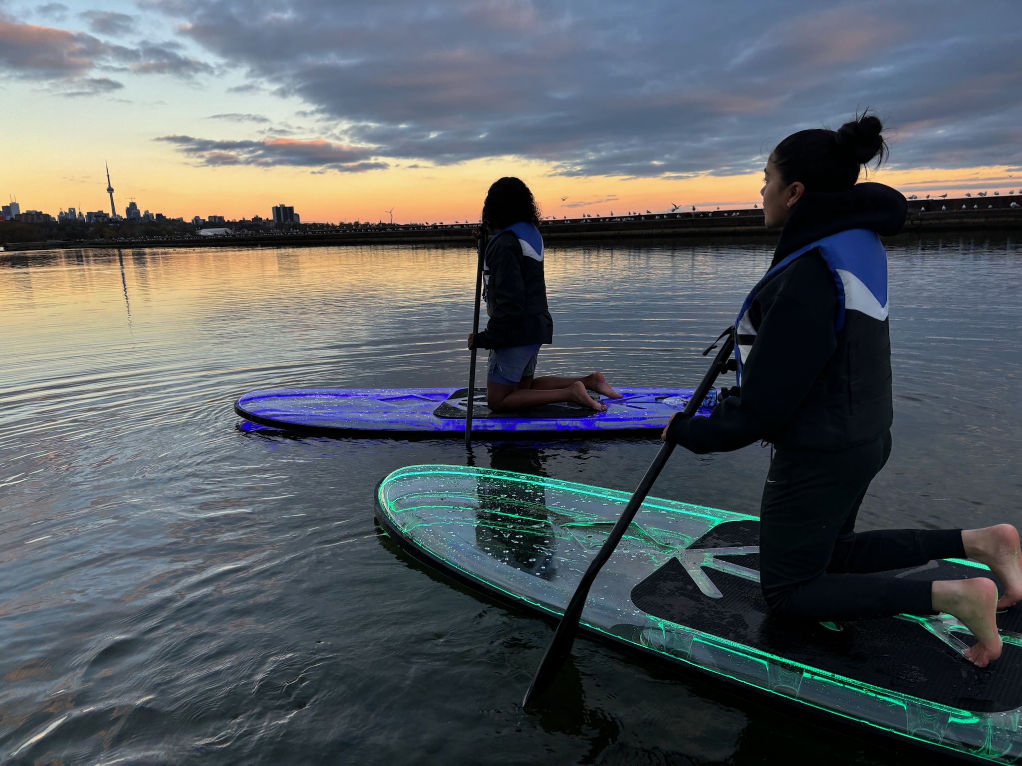 Two women glow in the dark paddleboarding on illuminated LED paddleboards at sunset on Lake Ontario with the Toronto skyline and CN Tower in the background