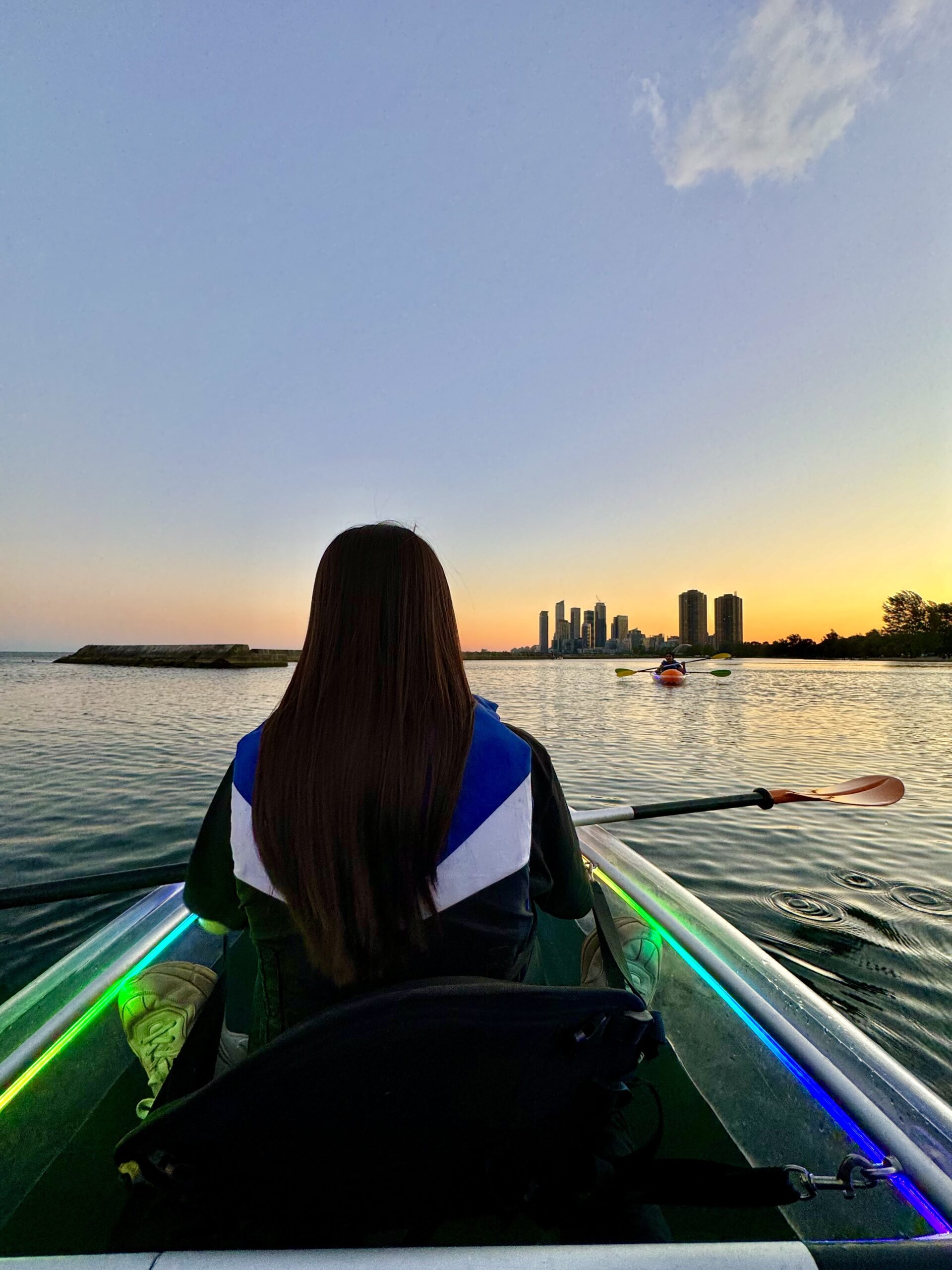 Woman paddling an illuminated clear kayak at sunset on Lake Ontario with the Toronto Humber Bay Arch Bridge skyline glowing on the horizon