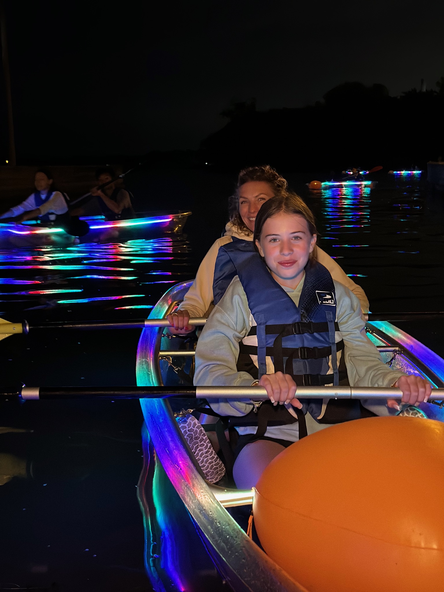 Mother and daughter smiling in a rainbow LED illuminated clear kayak at night in Toronto with a group of kayakers glowing behind them - Glow-In-The-Dark Kayaking is a fun family activity and things to do with kids in Toronto with Illuminated Escapes