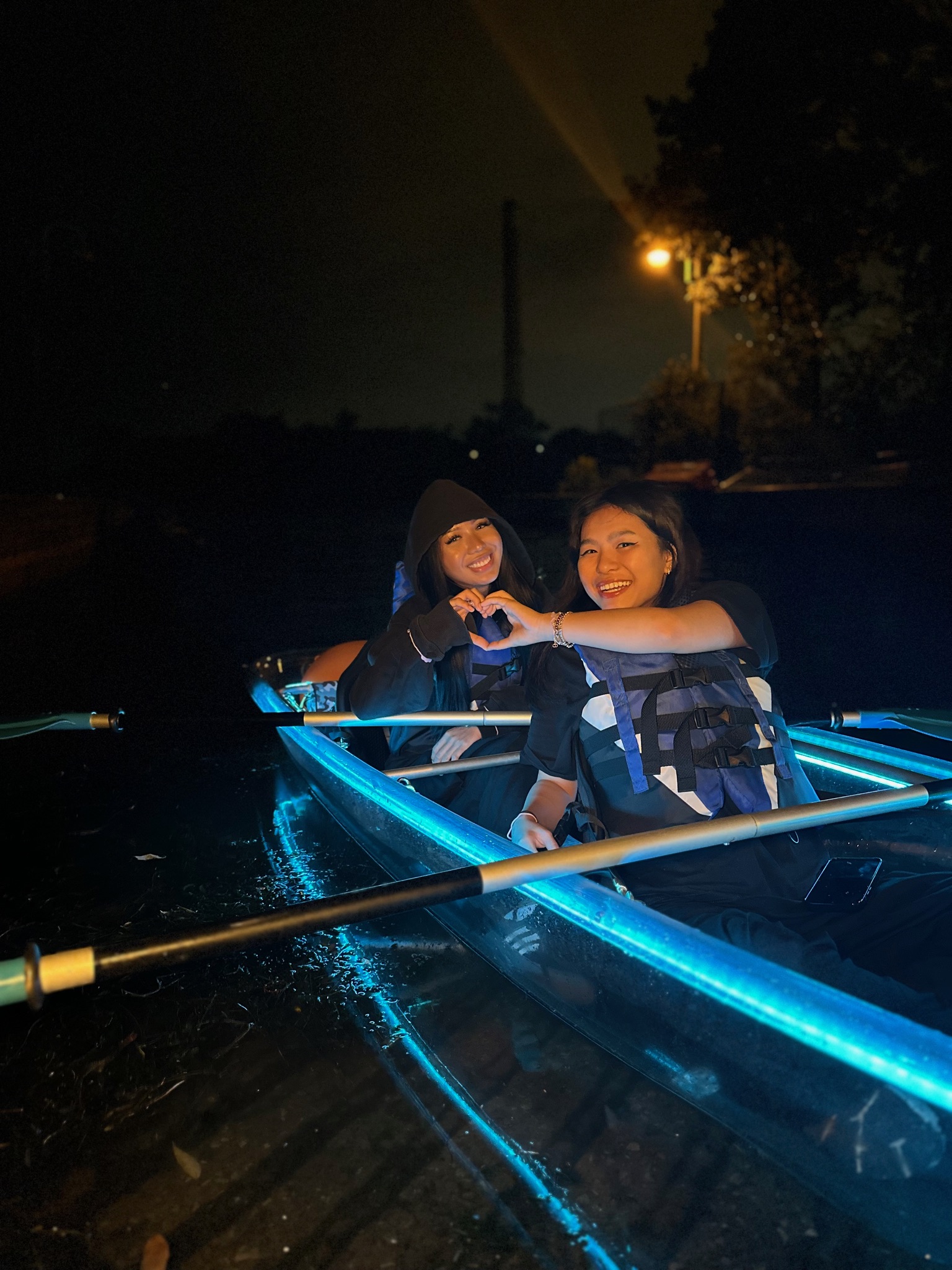 Two smiling friends making a heart shape with their hands in a blue LED illuminated clear kayak at night in Toronto - a fun things to do with friends summer night activity with Illuminated Escapes
