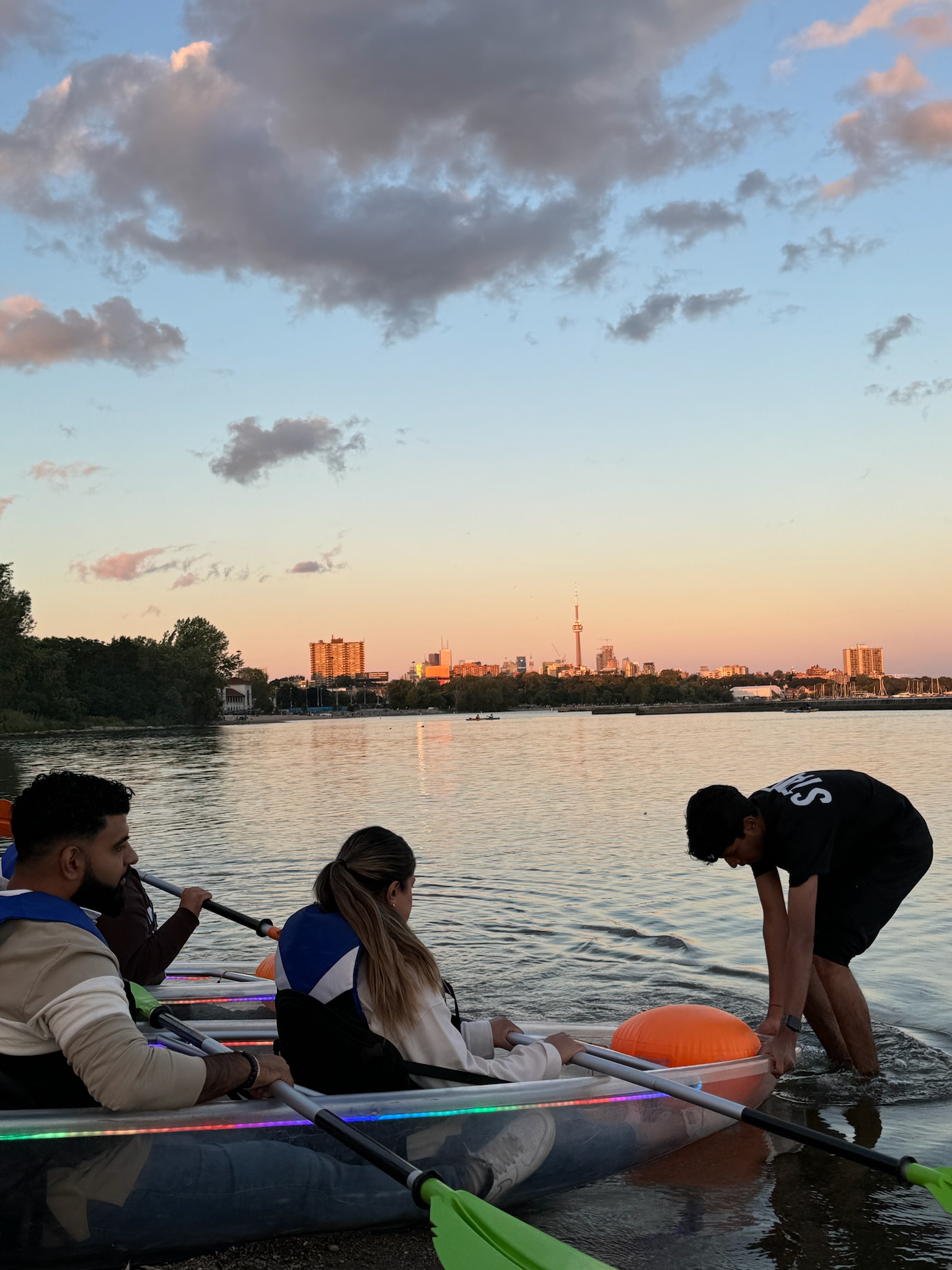 Group of friends launching an illuminated clear kayak at sunset on Lake Ontario with the Toronto skyline and CN Tower in the background. A unique summer activity with Illuminated Escapes
